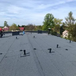 Wykończony dach pokryty papą termozgrzewalną z widocznymi odpowietrznikami i dwoma dekarzami pracującymi na dachu w słoneczny dzień.