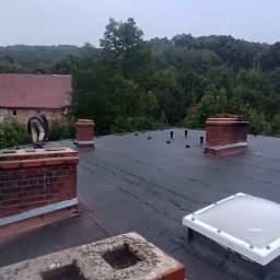 PAW-BUD usługi ogólnobudowlane - Czarny, płaski dach z kominami i świetlikiem. W tle budynek z czerwoną dachówką i zalesione wzgórze. Prace dekarskie w toku, widoczna sylwetka pracownika.