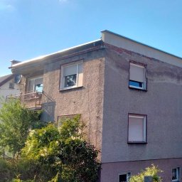 PAW-BUD usługi ogólnobudowlane - Dwupiętrowy budynek z szarą elewacją, widoczne ślady wcześniejszych prac murarskich na górnej kondygnacji, okna z roletami, fragment dachu i rynny, bujna zieleń w tle.