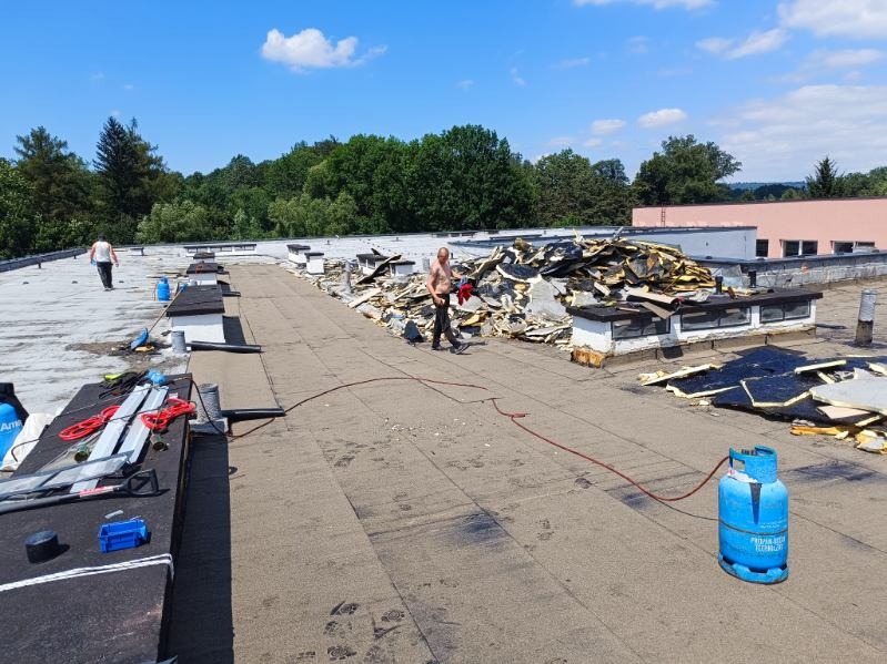 Demontaż starego pokrycia dachu płaskiego z widocznymi warstwami izolacji i papy, pracownik w trakcie pracy, butla gazowa na dachu, w tle drzewa i budynek.