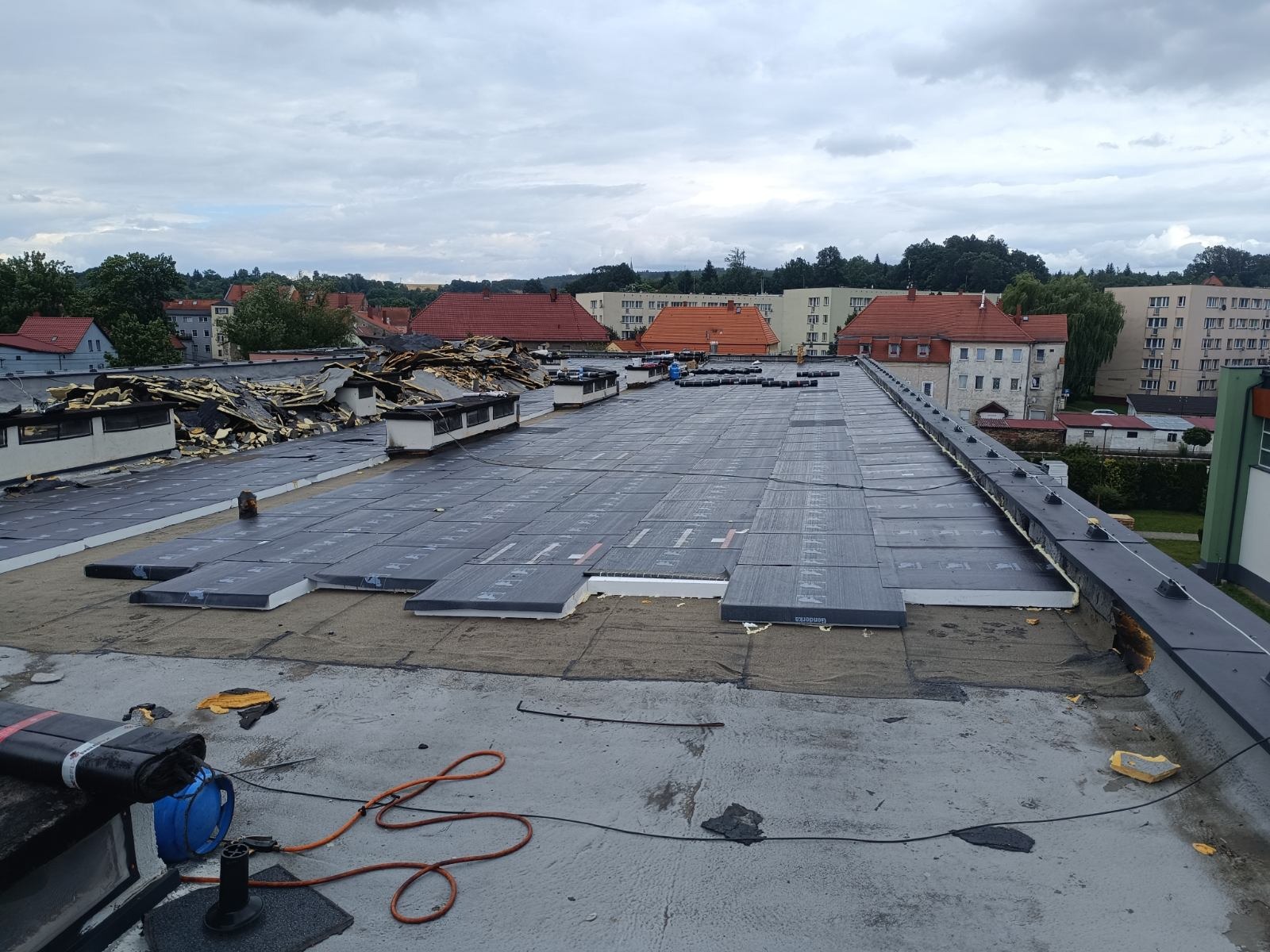 Prace na dachu płaskim budynku w trakcie wykonywania, widoczne warstwy izolacji termicznej i hydroizolacji, w tle zabudowa miejska i pochmurne niebo.
