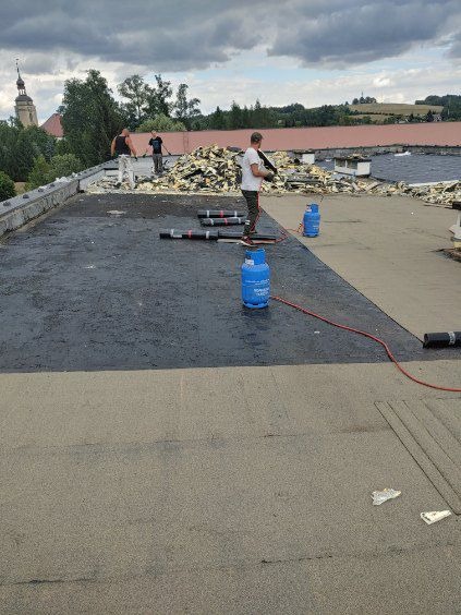 Prace dekarskie na płaskim dachu: robotnicy usuwają starą warstwę, przygotowując podłoże pod nowe pokrycie, widoczne butle z gazem i zwoje papy na tle miejskiego krajobrazu z wieżą kościelną.