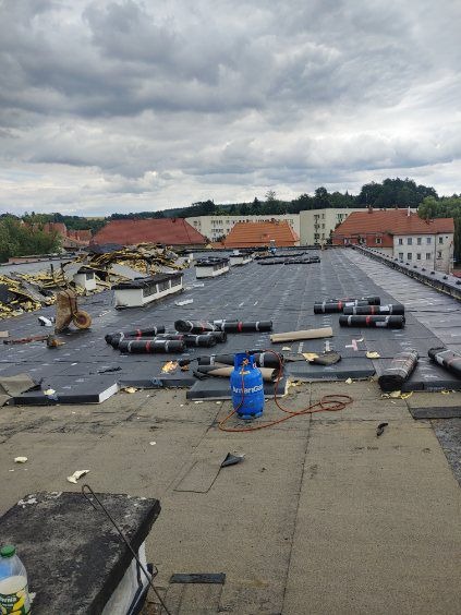 Rozległy płaski dach pokryty czarną papą termozgrzewalną w trakcie prac, widoczne rolki papy, butla gazowa z palnikiem, fragmenty starego pokrycia w oddali i wózek budowlany, w tle zabudowania...