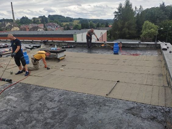 Pracownicy z palnikami na płaskim dachu podczas układania papy termozgrzewalnej, widoczne butle z gazem, budynki w tle.