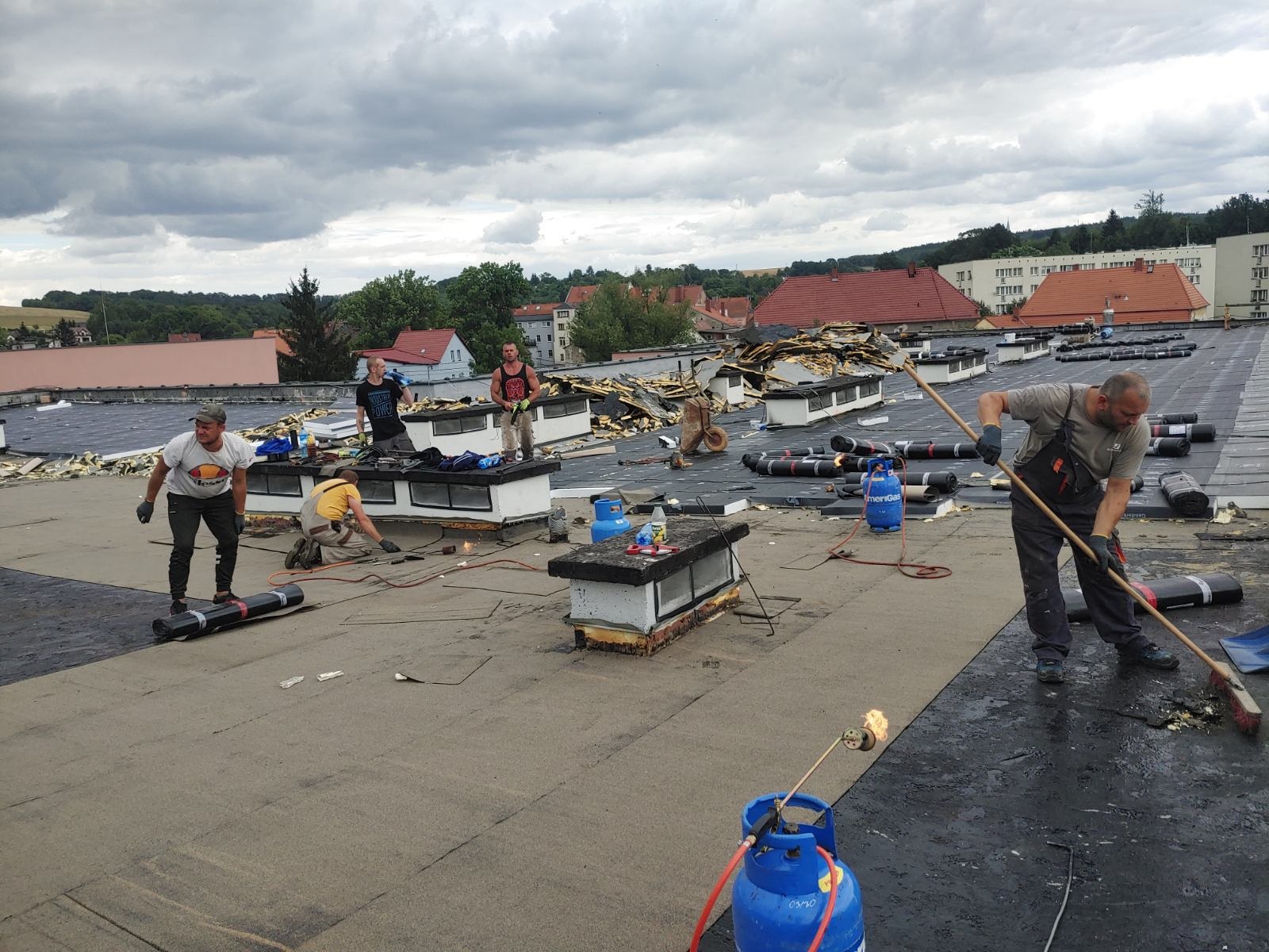 Ekipa dekarzy podczas prac na płaskim dachu budynku, z użyciem palnika i rolek papy termozgrzewalnej, widoczne butle gazowe i rozbiórka starej konstrukcji świetlika.