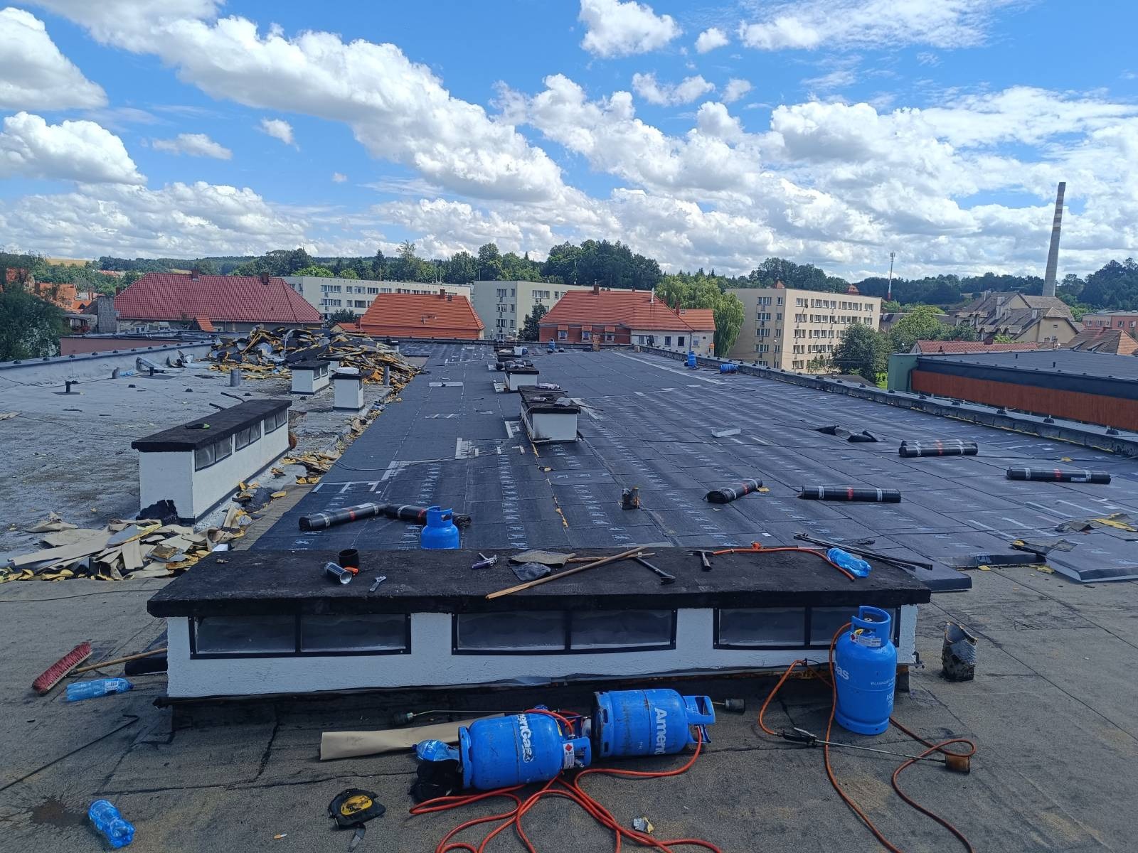Rozległy dach płaski w trakcie prac, widoczne fragmenty starego pokrycia, nowe czarne membrany, butle gazowe, narzędzia dekarskie i panorama miasta w tle pod błękitnym niebem z chmurami.