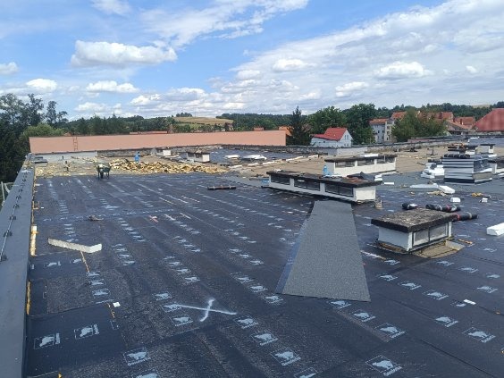 Duży, płaski dach pokryty czarną membraną z widocznymi oznaczeniami i częściowo usuniętą starą warstwą. W tle budynki mieszkalne i zieleń.