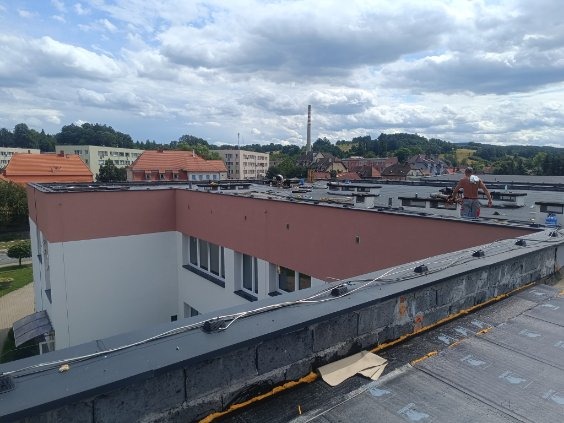 Prace na płaskim dachu budynku w trakcie realizacji, widoczny pracownik w oddali, materiały izolacyjne i elementy konstrukcyjne, w tle zabudowa miejska i komin.