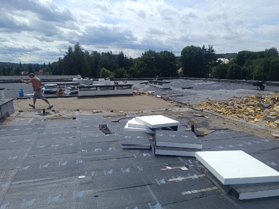 Prace dekarskie na płaskim dachu w trakcie realizacji: widoczne fragmenty starego pokrycia, nowe płyty izolacyjne styropianowe, dekarz z butlą gazową w tle, krajobraz zalesiony w oddali.