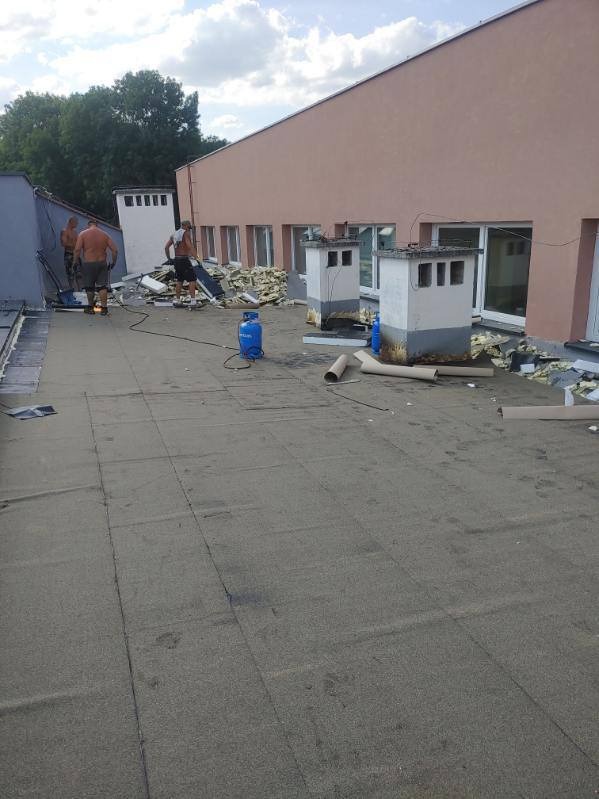 Ekipa dekarzy podczas prac na płaskim dachu budynku z kominami, widoczne butle gazowe i resztki materiałów budowlanych.