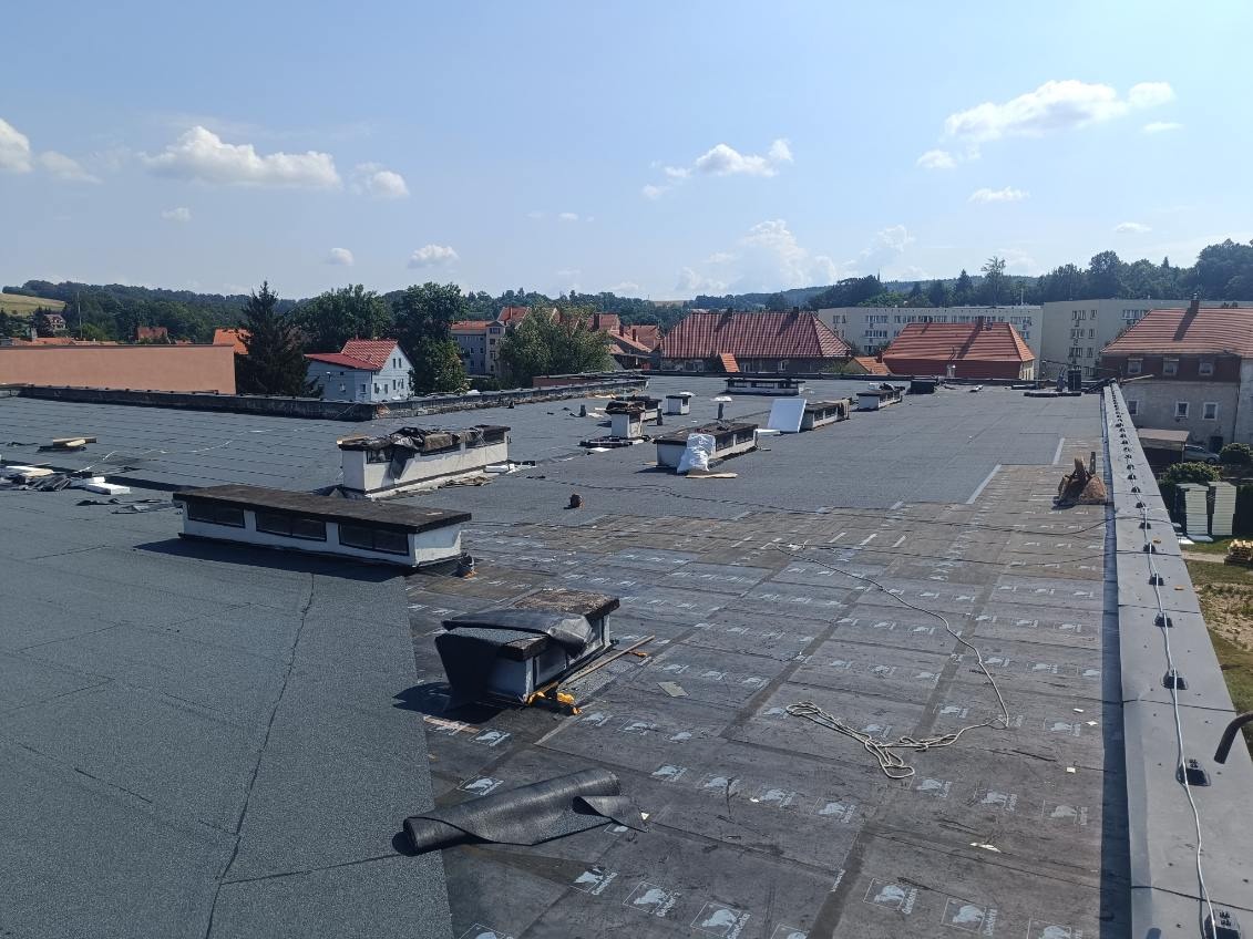 Rozległy, płaski dach pokryty papą w trakcie prac, widoczne elementy wentylacyjne, fragmenty starego pokrycia oraz zwinięta rolka papy. W tle zabudowa miejska i błękitne niebo.
