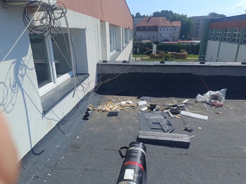Prace na płaskim dachu budynku z widocznymi elementami starej izolacji, narzędziami i zwisającymi kablami, w tle zabudowa miejska.