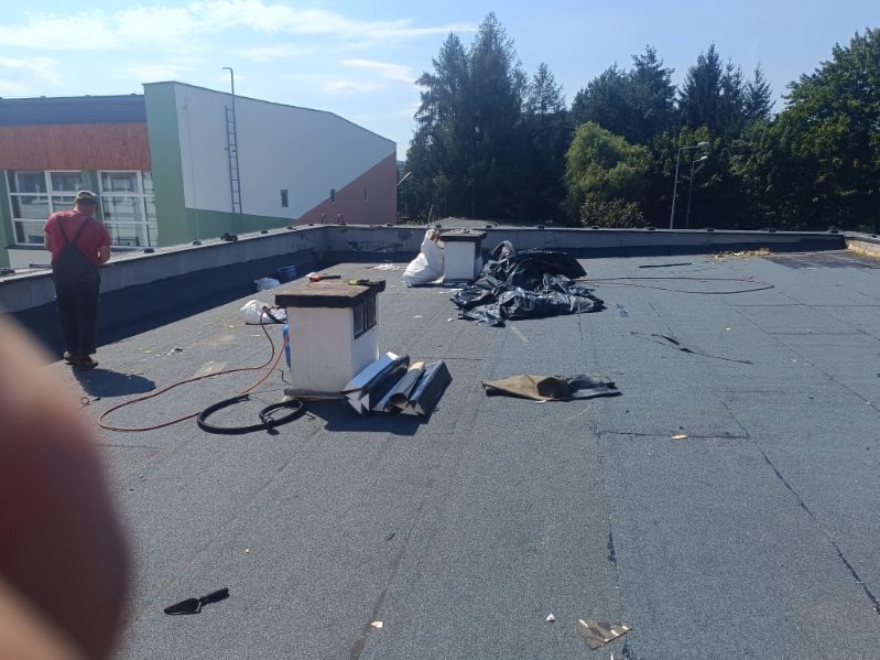 Prace na płaskim dachu pokrytym papą, widoczne kominki wentylacyjne, przygotowane elementy blacharskie, narzędzia i pracownik w oddali.