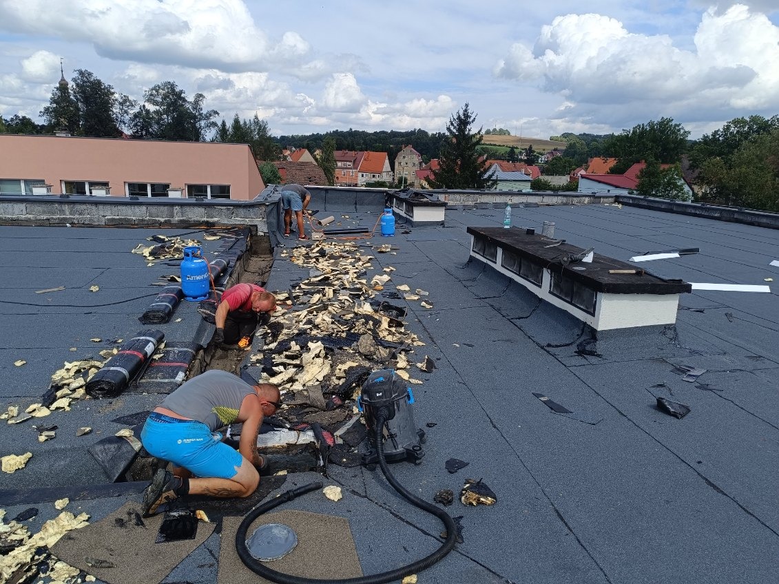 Ekipa dekarzy podczas prac na płaskim dachu, usuwanie starej papy i przygotowanie podłoża pod nowe pokrycie, widoczne butle z gazem, odkurzacz przemysłowy i narzędzia, w tle zabudowa miejska...