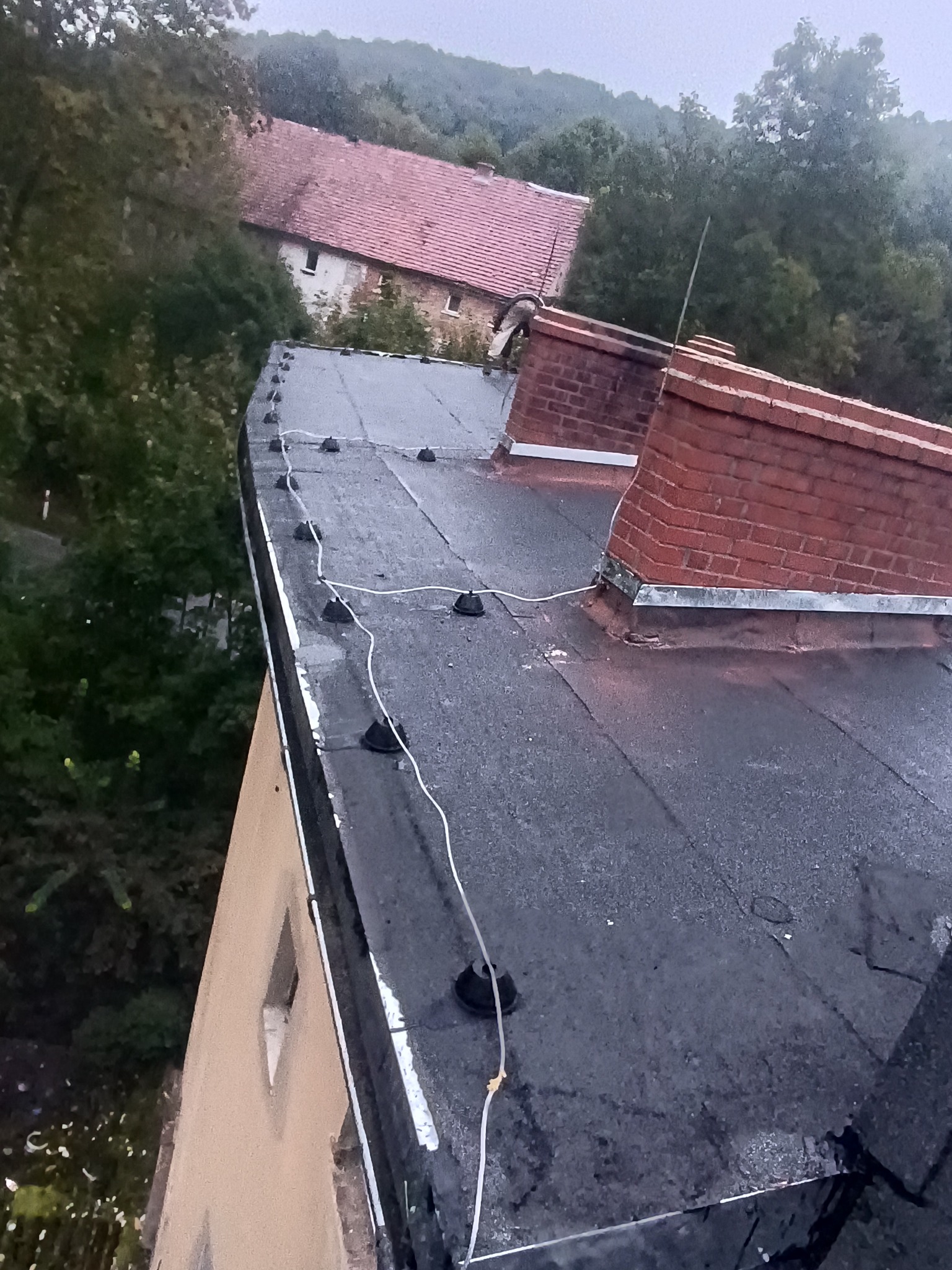 Czarny, płaski dach z instalacją odgromową, kominem z cegły i widokiem na budynek z czerwonym dachem w tle, sfotografowany z góry pod kątem.