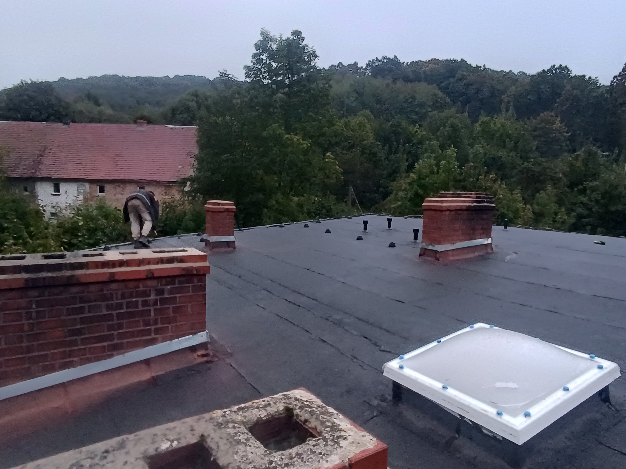 Czarny, płaski dach z kominami i świetlikiem. W tle budynek z czerwoną dachówką i zalesione wzgórze. Prace dekarskie w toku, widoczna sylwetka pracownika.