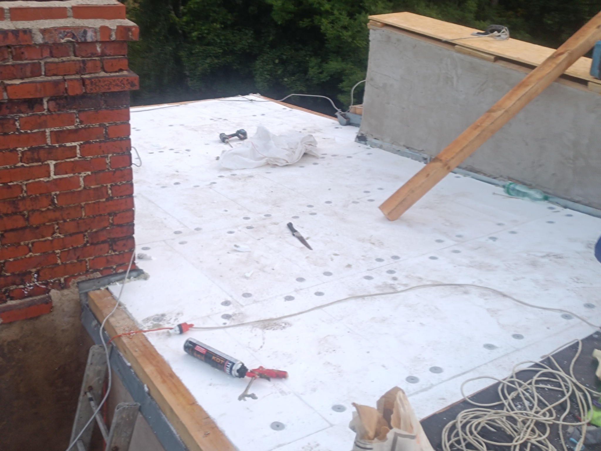 Dach w trakcie ocieplania: białe płyty izolacyjne z widocznymi mocowaniami, komin z czerwonej cegły, narzędzia i materiały budowlane. Prace dekarskie w toku.