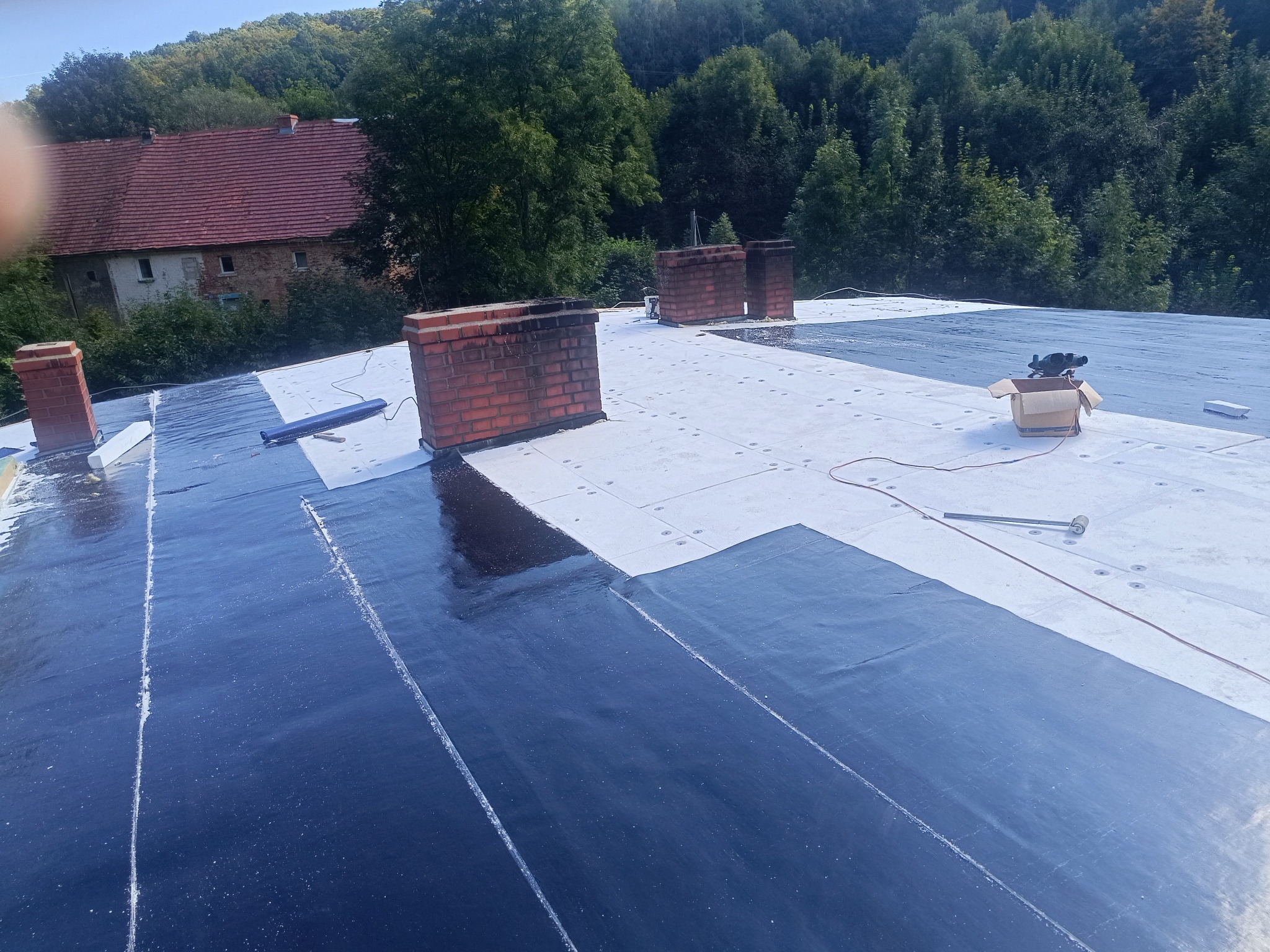 Dach z częściowo położoną, czarną membraną dachową, widoczne kominy z czerwonej cegły, narzędzia i budynek w tle, otoczony zielenią.