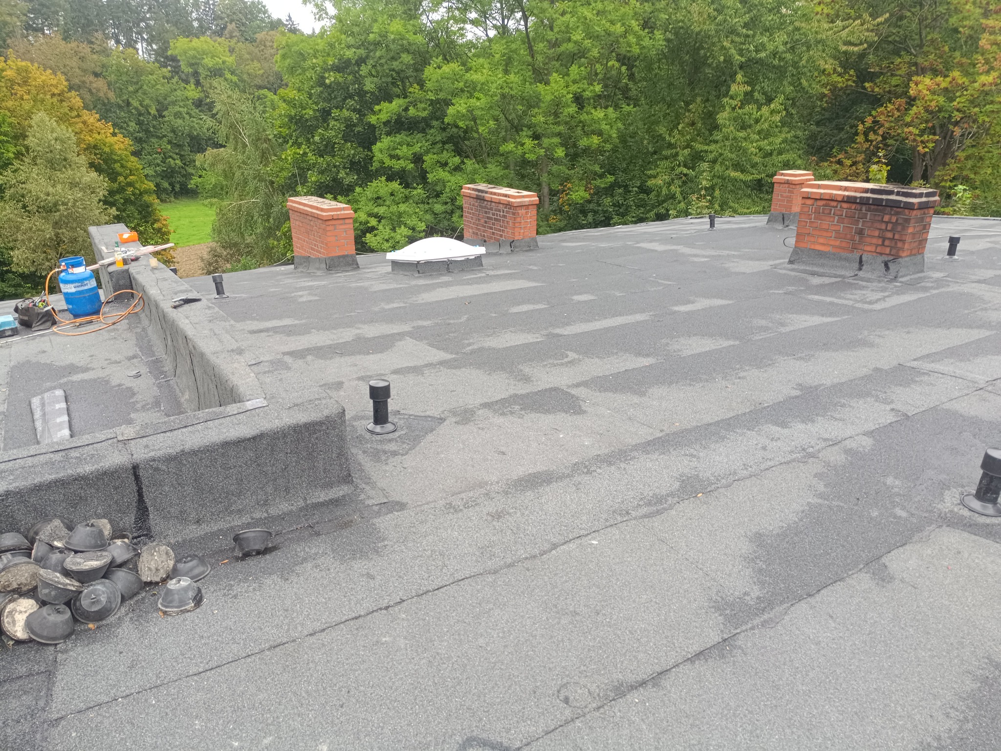 Dach pokryty papą z kominami z cegły i elementami wentylacyjnymi. Na dachu leżą narzędzia i akcesoria dekarskie. Widok z góry na tle zieleni.