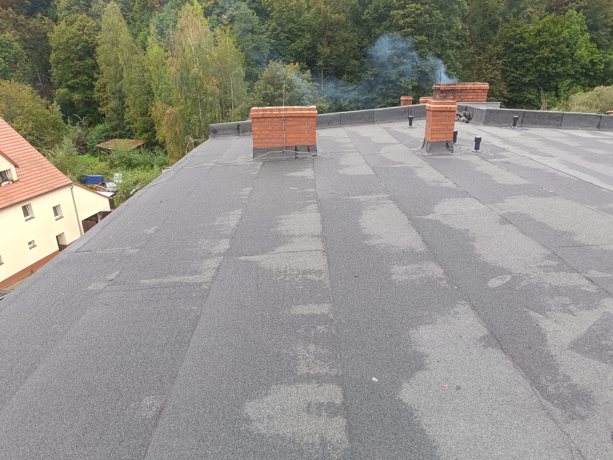 Widok z góry na płaski dach pokryty papą z ceglanymi kominami. W tle drzewa i fragment budynku z czerwoną dachówką.