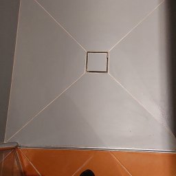 Mój Market Avetik Vanetsyan - Szara, kwadratowa kabina prysznicowa z odpływem liniowym, pomarańczowe płytki podłogowe i fragment stopy w czarnej skarpetce.