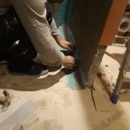 Mój Market Avetik Vanetsyan - Pracownik w klapkach aplikuje folię hydroizolacyjną na ścianę w rogu pomieszczenia przed pracami glazurniczymi, widoczny fragment drabiny i młotek na podłodze.