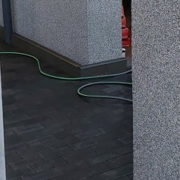 Mój Market Avetik Vanetsyan - Ułożona, ciemna kostka brukowa wzdłuż szarej ściany, z widocznym fragmentem zielonego węża ogrodowego i stosem czerwonych kostek w tle.