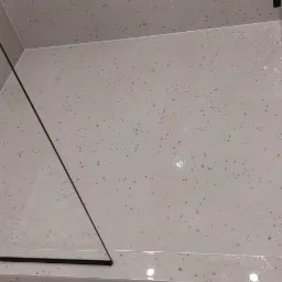 Wykończony brodzik kabiny prysznicowej z odpływem liniowym, wyłożony jasnymi płytkami w drobny deseń, częściowo osłonięty szklaną taflą.