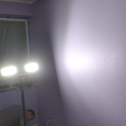 Mój Market Avetik Vanetsyan - Malowanie fioletowej ściany w pokoju, widoczna lampa halogenowa na statywie oświetlająca fragment ściany, mężczyzna szpachluje podłogę przy ścianie.