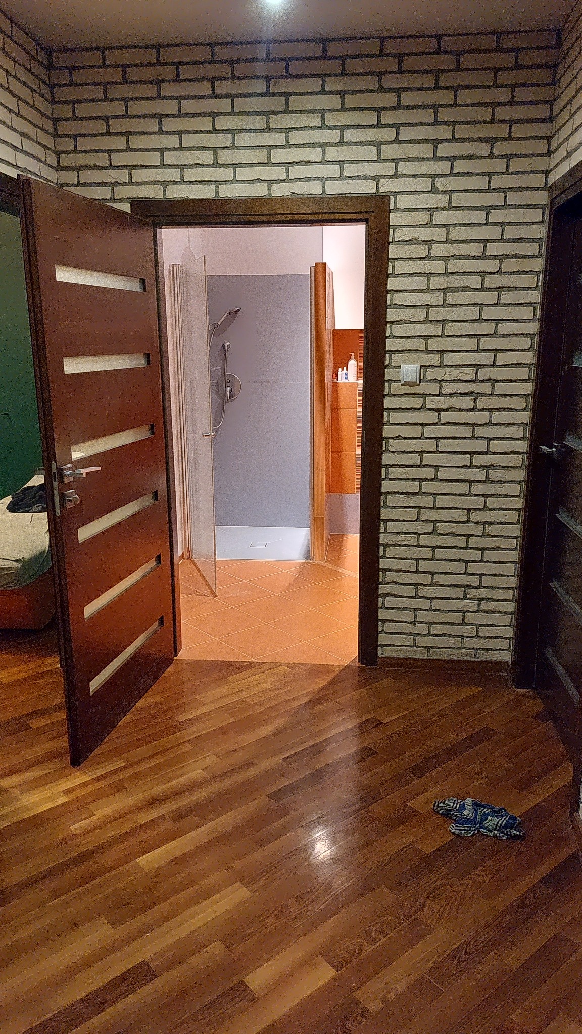 Otwarta łazienka z kabiną prysznicową, widok z korytarza na drewnianą podłogę i ścianę z cegieł dekoracyjnych, z fragmentem pozostawionej szmatki na podłodze.