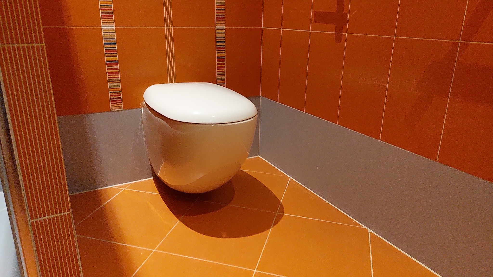 Wisząca muszla klozetowa z białą deską na tle pomarańczowych płytek z dekoracyjnymi paskami, szara lamperia, pomarańczowa podłoga z białymi fugami.