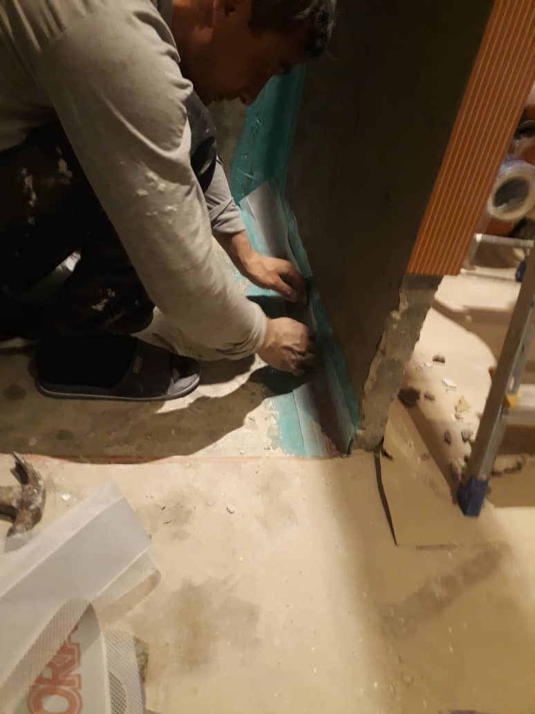 Pracownik w klapkach aplikuje folię hydroizolacyjną na ścianę w rogu pomieszczenia przed pracami glazurniczymi, widoczny fragment drabiny i młotek na podłodze.