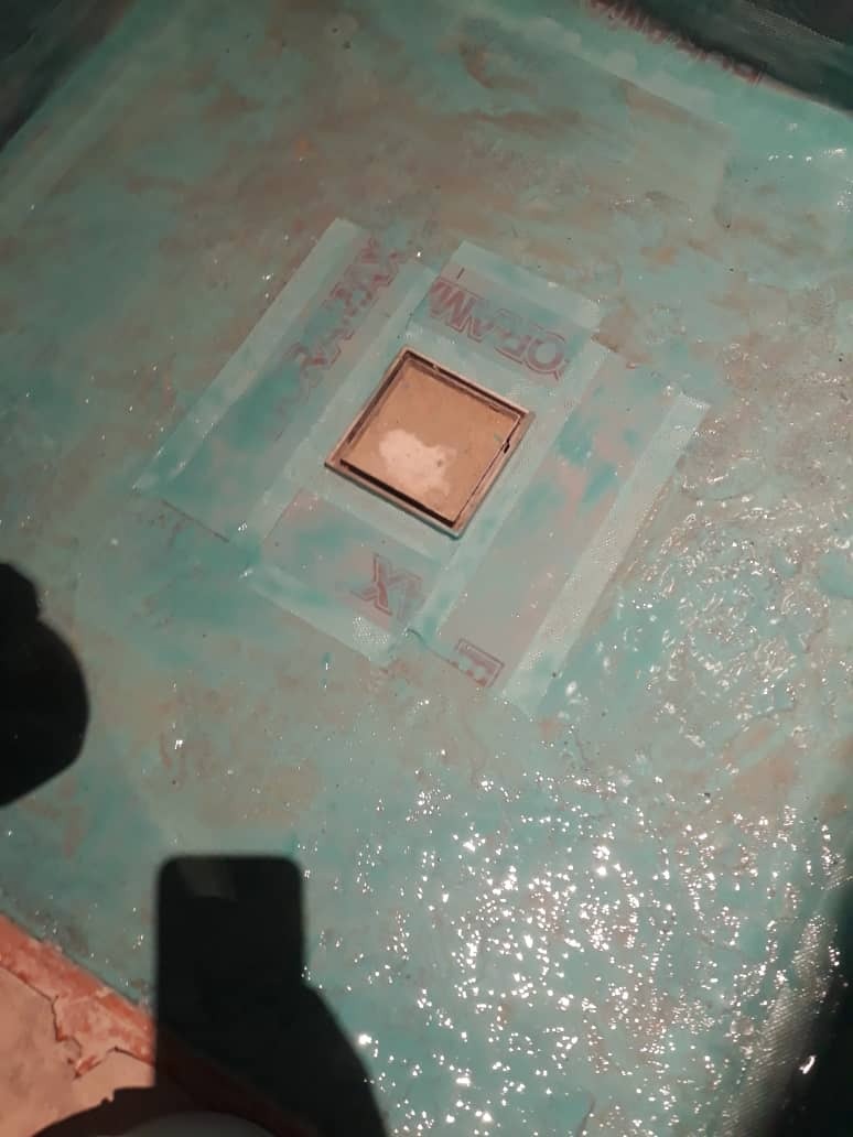 Hydroizolacja podłoża w brodziku prysznicowym, zabezpieczona taśmami uszczelniającymi wokół odpływu liniowego, widoczne mokre podłoże pokryte warstwą hydroizolacji w odcieniach zieleni i błękitu.