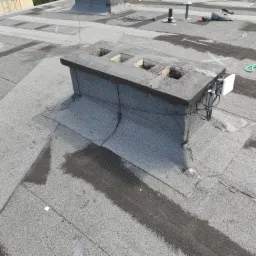 Płaski dach pokryty papą z widocznym kominem z trzema otworami, anteną i śladami wilgoci.