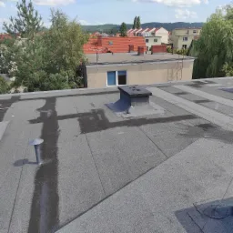 Płaski dach pokryty papą w Rumi, z widocznymi śladami po deszczu wokół komina i wentylacji, na tle zabudowy miejskiej.