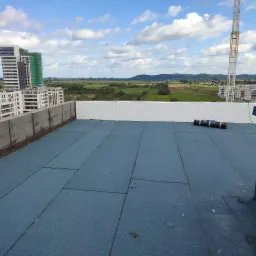 Widok z góry na płaski dach pokryty niebieską membraną, z rolką papy w tle i panoramą miasta z budynkami i żurawiem budowlanym w oddali.