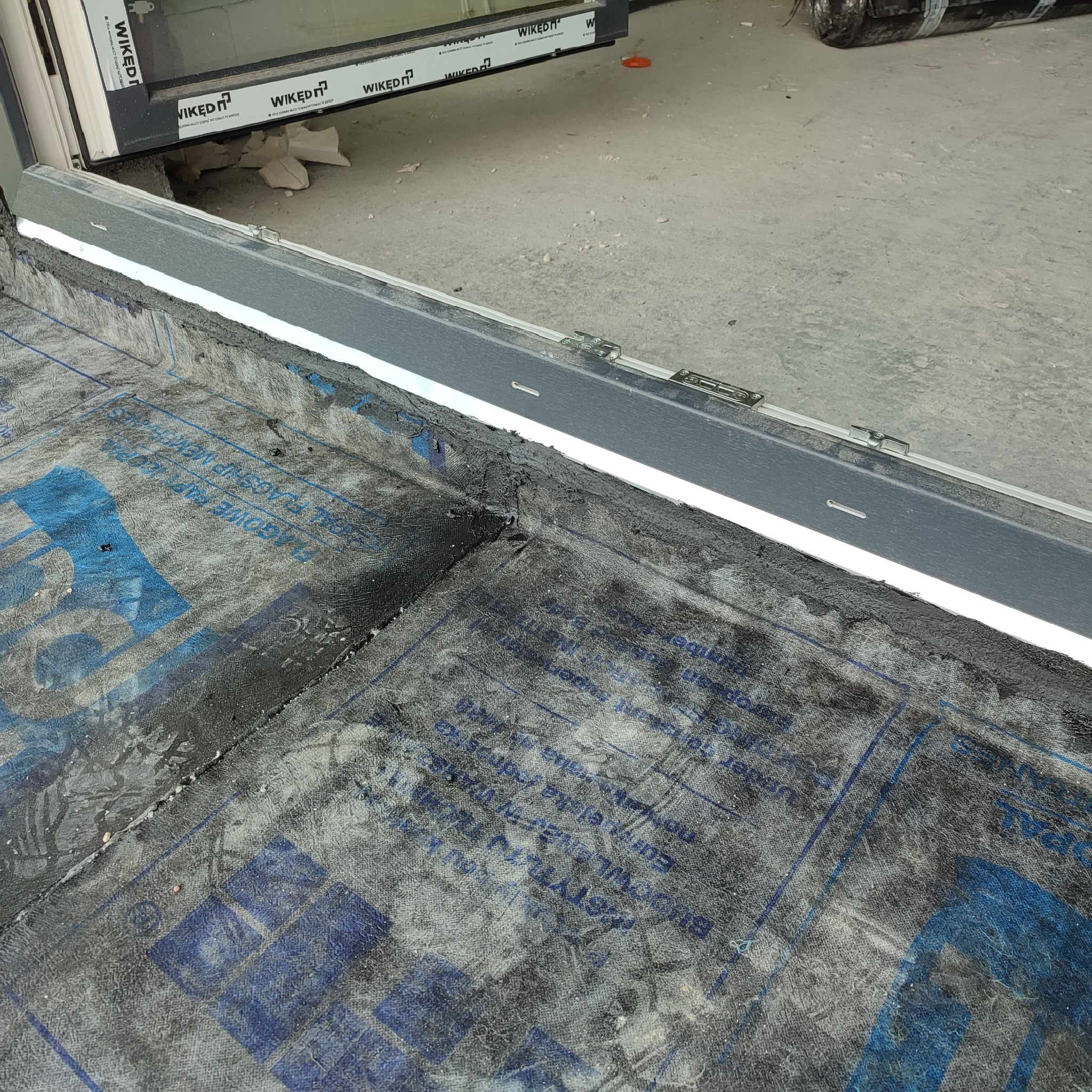 Fragment dachu z widoczną hydroizolacją z papy podkładowej z niebieskim nadrukiem, przytwierdzoną do obróbki blacharskiej okna dachowego.