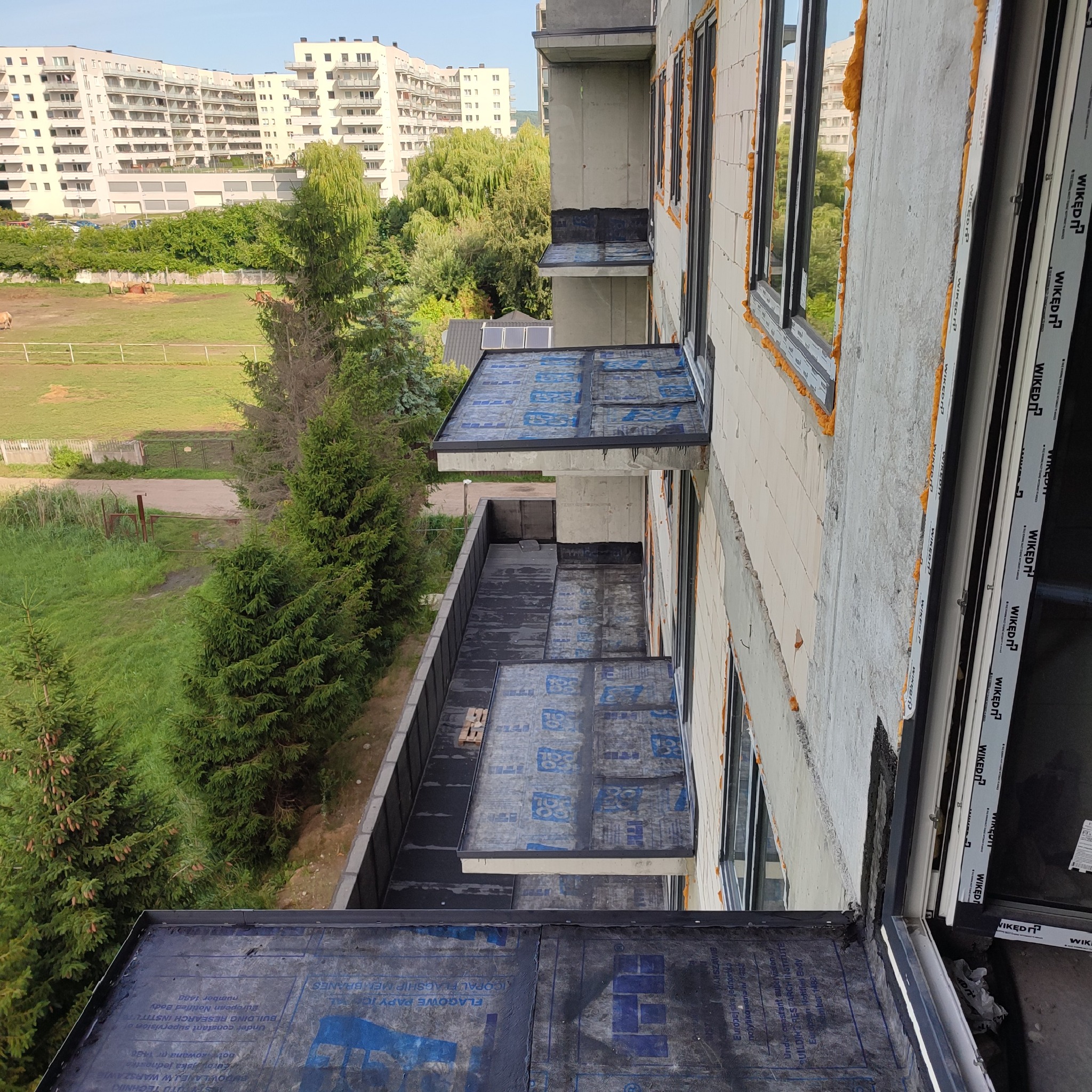 Budynek w trakcie budowy, widok z góry na balkony pokryte papą, okna z pianką montażową, w tle zieleń i budynki mieszkalne.