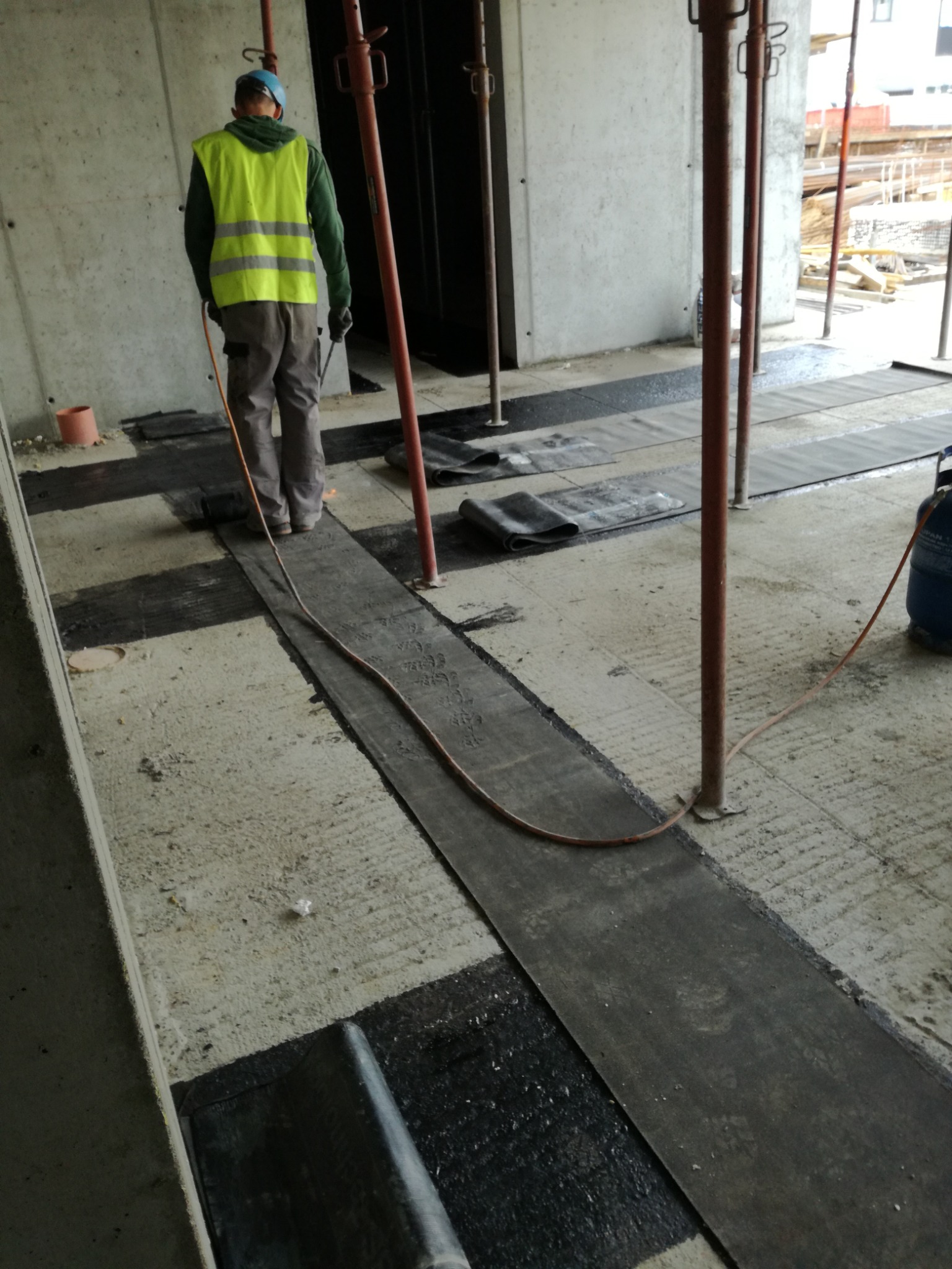 Pracownik w kamizelce odblaskowej układa pasy bitumiczne na betonowej powierzchni, używając palnika; widoczne stalowe podpory i butla z gazem.