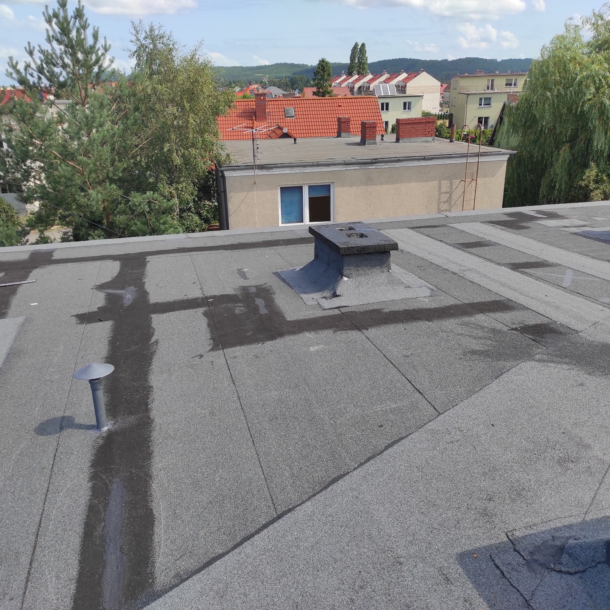 Płaski dach pokryty papą w Rumi, z widocznymi śladami po deszczu wokół komina i wentylacji, na tle zabudowy miejskiej.