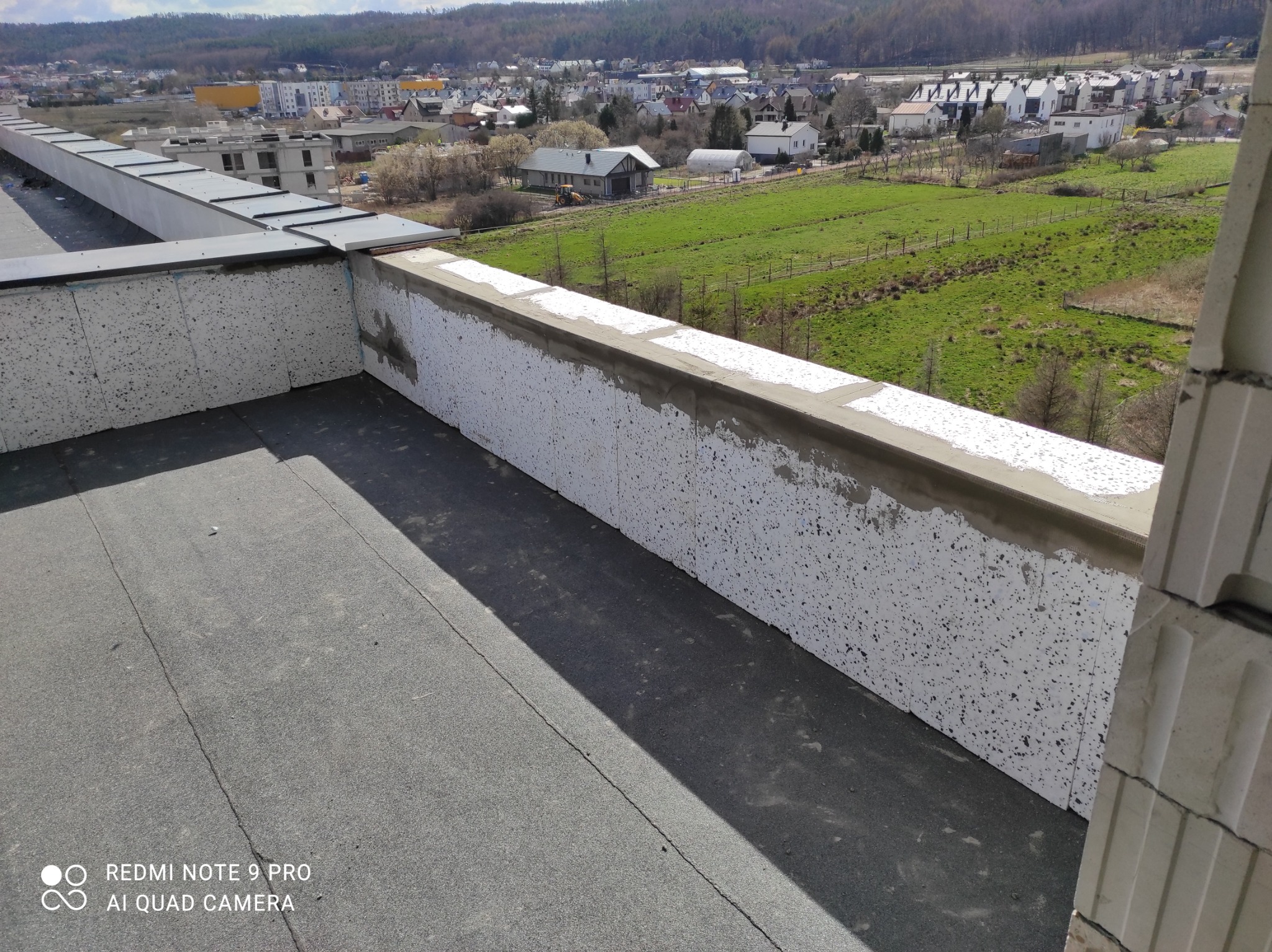 Szary dach z papy termozgrzewalnej, zabezpieczony styropianem i betonową obróbką, z widokiem na okoliczną zabudowę i zalesione wzgórza w tle.