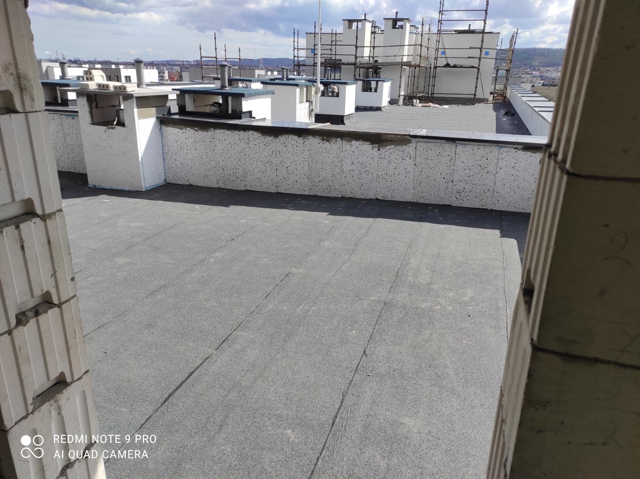 Widok z góry na płaski dach pokryty papą, z kominami wentylacyjnymi i rusztowaniami w tle, widziany zza fragmentu muru z bloczków.
