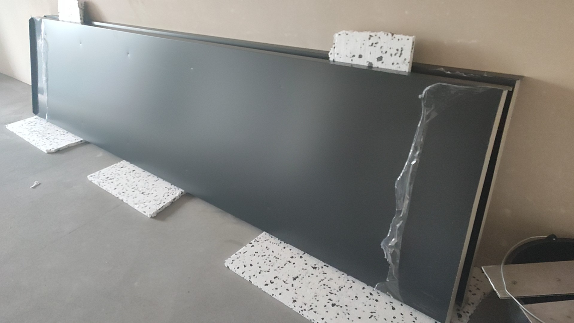 Szary panel dachowy z metalowym wykończeniem, zabezpieczony folią ochronną na krawędzi, podparty na białych płytkach styropianowych na szarym podłożu, przygotowany do montażu.