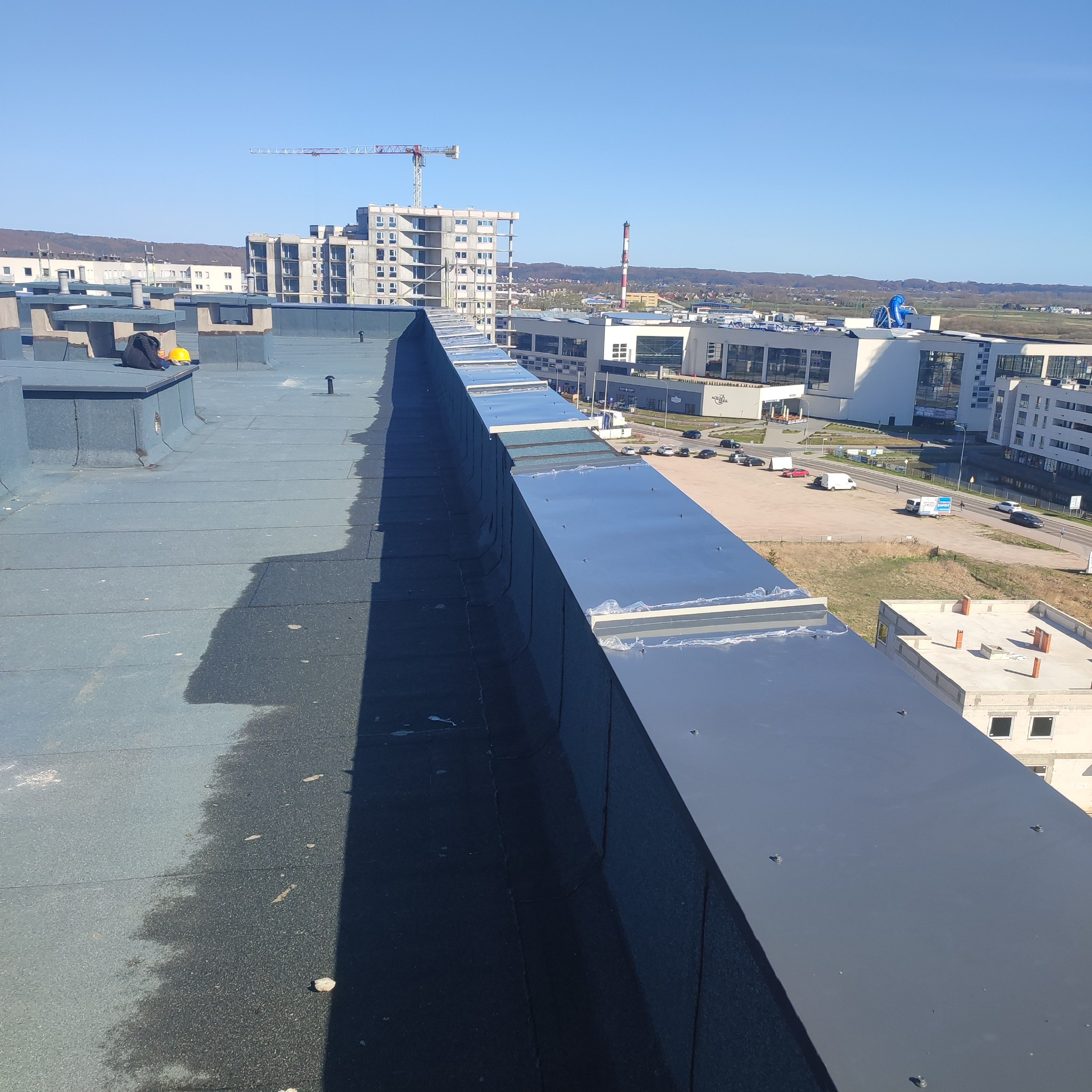 Widok z góry na płaski dach pokryty papą termozgrzewalną, z widocznymi obróbkami blacharskimi na krawędziach, w tle zabudowa miejska i budowa z dźwigiem.
