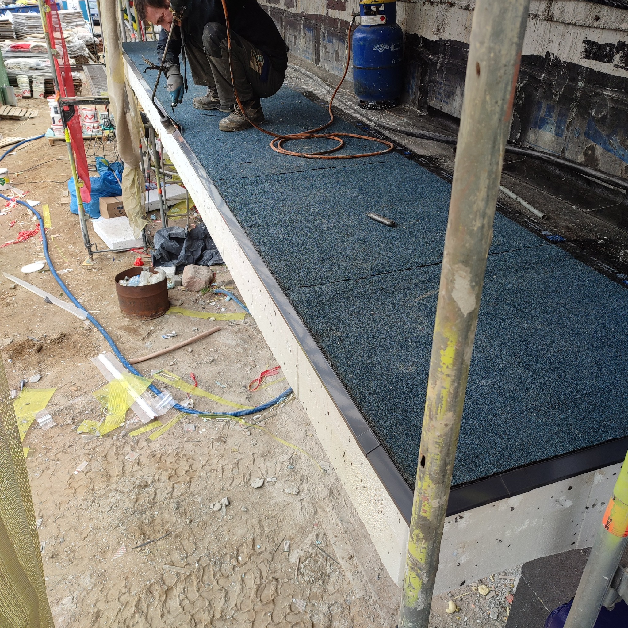 Pracownik w trakcie uszczelniania połączeń papy termozgrzewalnej na dachu płaskim za pomocą palnika gazowego; widoczna butla gazowa, narzędzia i rusztowanie.