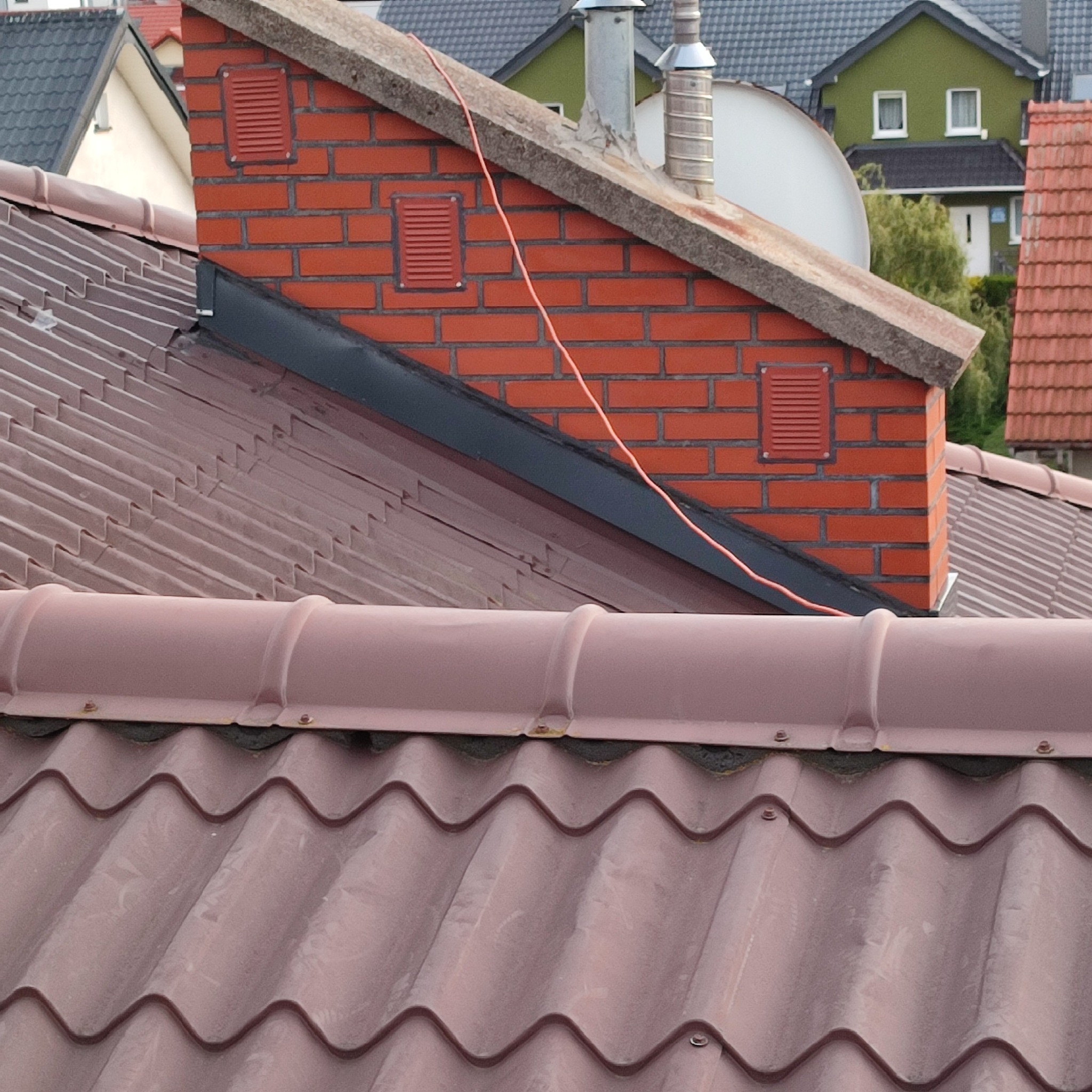 Czerwony ceglany komin z kratkami wentylacyjnymi, wystający ponad brązowy dach z blachy falistej na tle zabudowy jednorodzinnej.