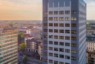 Wysoki budynek biurowy z szarą elewacją w promieniach zachodzącego słońca, widoczny z góry na tle panoramy miasta.