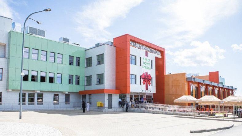 Nowoczesny budynek szkoły podstawowej z elewacją w odcieniach zieleni, bieli i pomarańczy, ozdobiony dużą czerwoną kokardą nad wejściem, z widocznymi osobami wchodzącymi do budynku.