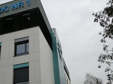 Nowoczesna elewacja budynku biurowego z panelami w odcieniach szarości i turkusu, widoczne okna z żaluzjami zewnętrznymi i fragment nazwy budynku na dachu, pochmurne niebo w tle.