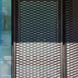 Metalowa siatka o strukturze plastra miodu, zamontowana na elewacji budynku, tworząca ażurową przesłonę okienną. Widoczna szklana ściana za siatką, częściowo zasłonięta folią ochronną.